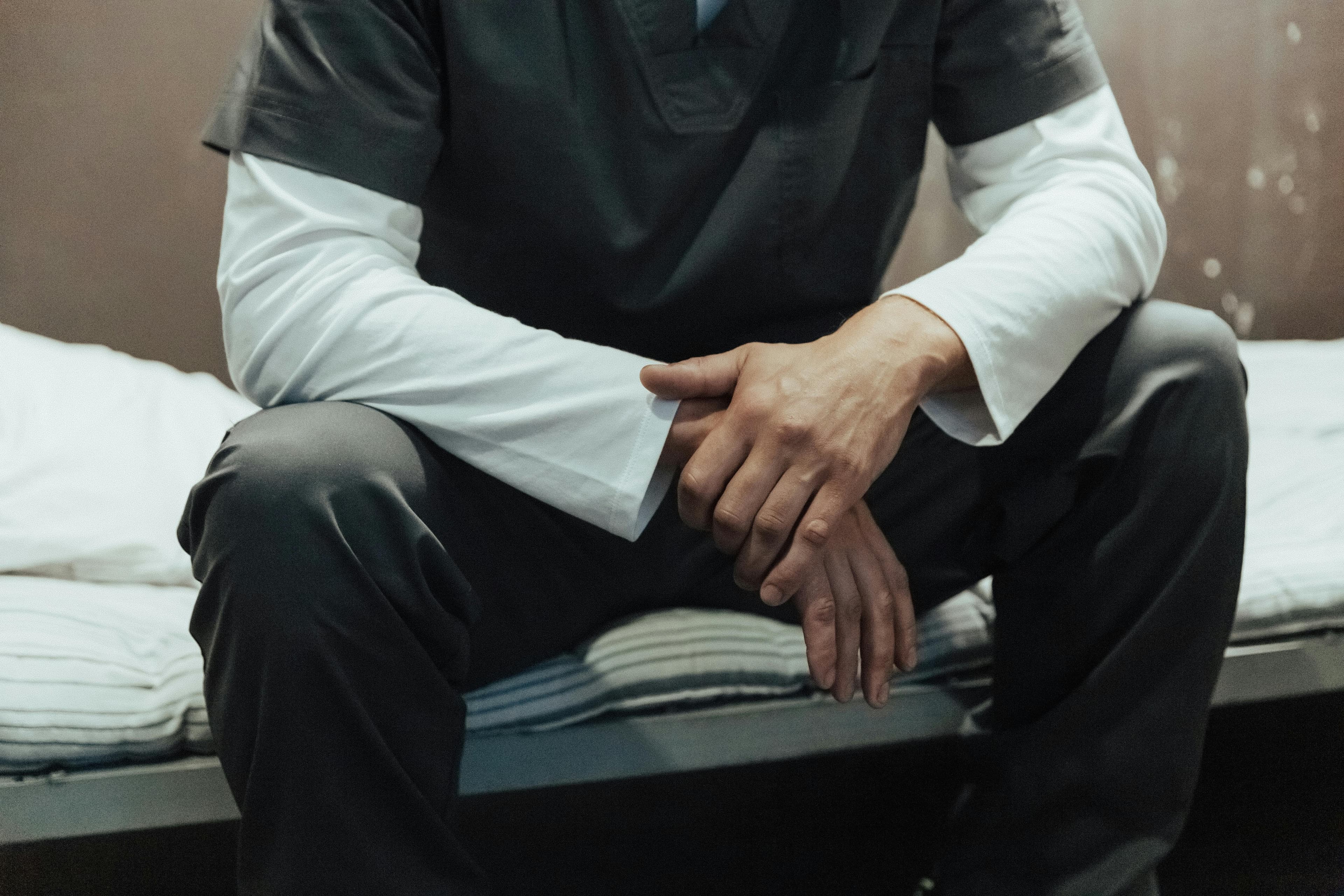 A nurse in scrubs sitting on the edge of a hospital bed, hands clasped, at the end of a shift.